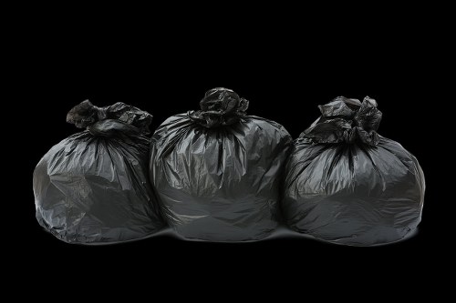 Three large black rubbish bags made of thick, glossy plastic material are lined up on a flat surface against a black background. The bags are tightly knotted at the top, with the middle bag positioned slightly in front of the others, and the two on either side leaning slightly inward. The plastic bags exhibit a shiny finish with visible folds and creases, and their irregular shapes suggest they are filled with miscellaneous waste. The environment appears to be a controlled, indoor setting, possibly part of a waste collection or disposal process, with no other objects or context visible. The focus on the rubbish bags aligns with the service provided by Commercial Waste Removal Lambeth, highlighting their capability in handling waste management for commercial clients in the Lambeth area, near postcode SW9 or SW2.