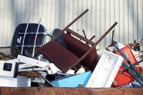 A large pile of various discarded household items and rubbish sits outdoors against a metal wall, including a maroon upholstered chair with wooden legs, a white plastic window frame, a blue plastic container, a red and black plastic chair, and a black metal shopping basket. The objects are stacked haphazardly, with some items resting on top of others, indicating they are awaiting removal or disposal. The scene appears to be on a driveway or yard, with natural daylight illuminating the clutter. The presence of different materials such as plastic, wood, and metal reflects common waste items collected during rubbish clearance services provided by Commercial Waste Removal Lambeth, serving the Lambeth area post code region. The overall image captures a typical scene of household waste awaiting collection, emphasizing the need for professional rubbish removal in the local community.