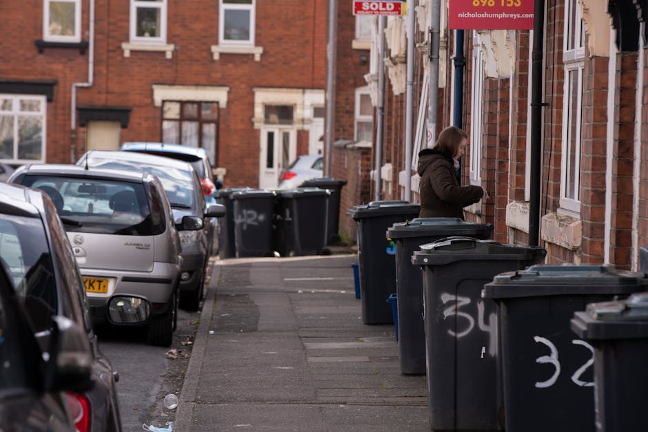 A middle-aged man with dark hair, dressed in a black t-shirt and dark trousers, is seen disposing of rubbish into a stainless steel public litter bin on a paved pathway. The man is holding a large whi